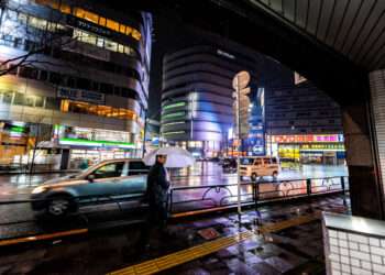 Shinjuku, Japan - April 1, 2019: Exterior of station in downtown Tokyo city with neon bright lights signs reflection at night in rain with man businessman under umbrella