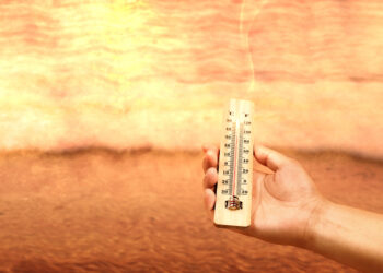 Hand holding thermometer with high temperature on the beach with glowing sun background. Heatwave concept
